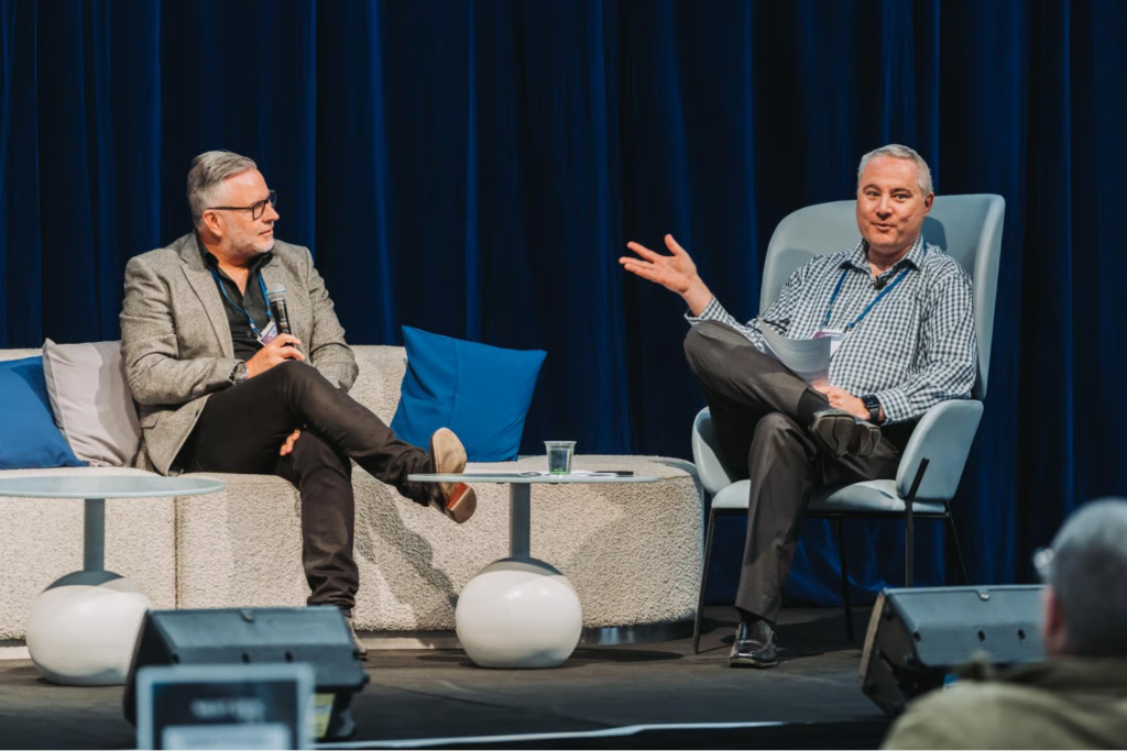Two men seated on stage in conversation, one holding a microphone, at a conference.