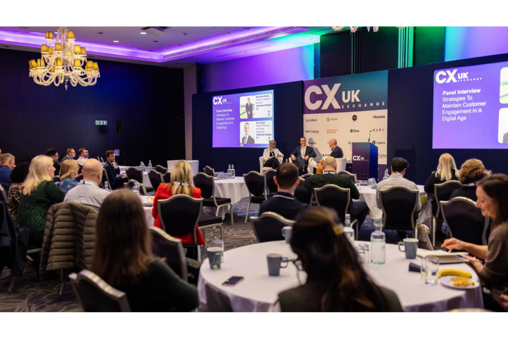 Panel interview on stage at CX UK Exchange conference with seated audience at round tables.